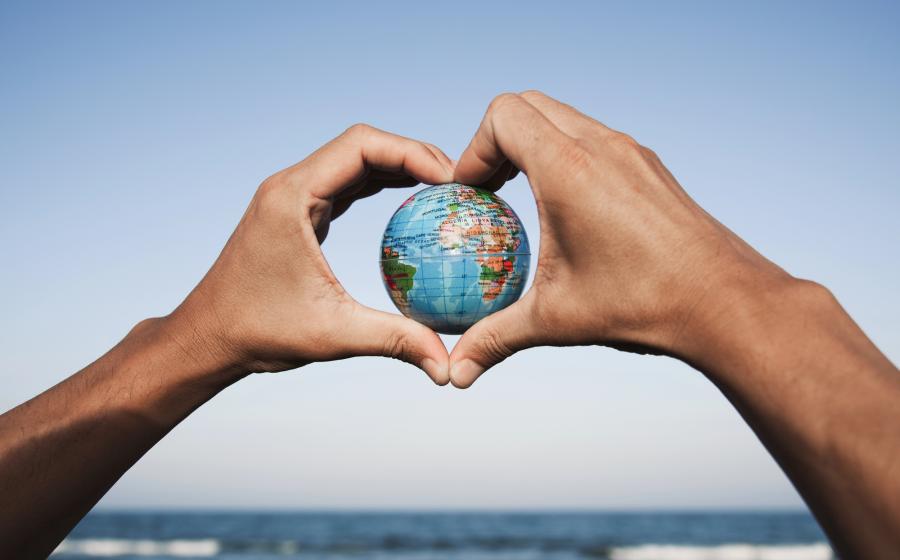 Hands in a heart shape holding a small globe, with sky and waves in the background