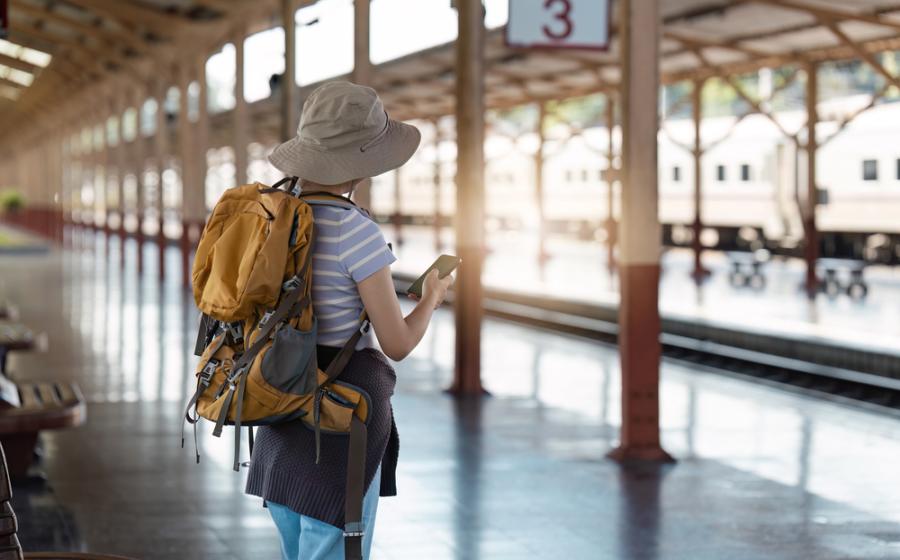 A solo traveler with a backpack and hat stands at a train station platform, checking their phone for travel updates