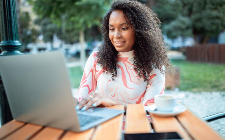 A woman with curly hair focuses on her laptop while sitting at a wooden table. She enjoys a cup of coffee and has a smartphone nearby, surrounded by greenery