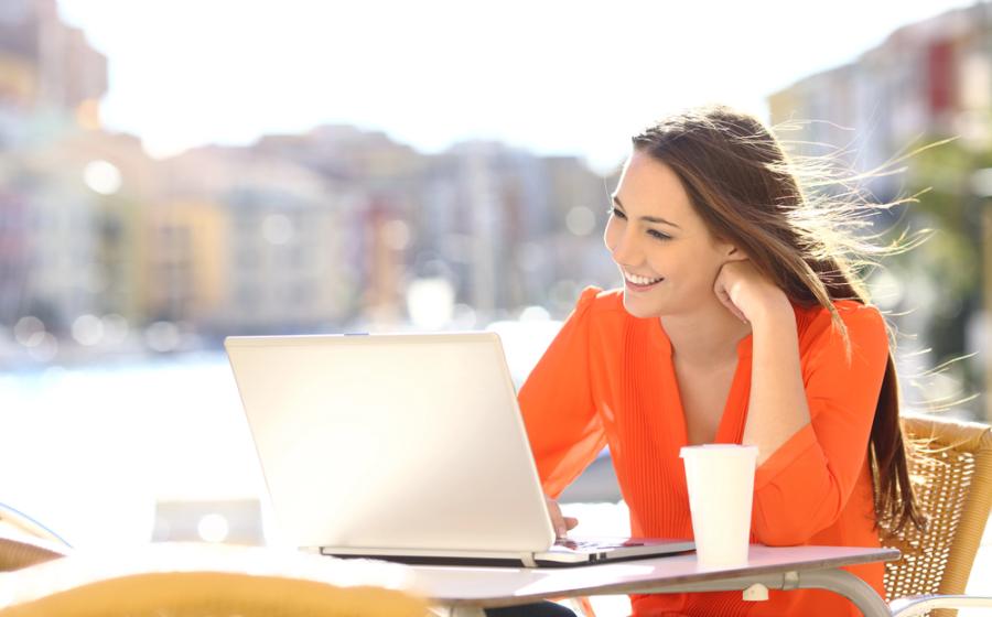 Happy lady in a coffee shop terrace uses a laptop in a coast town street